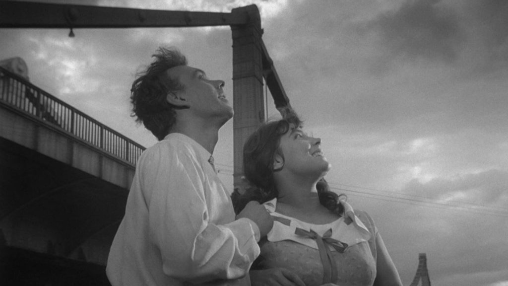 A man and woman look up admiringly at the sky with a large bridge behind them in the film "The Cranes Are Flying."