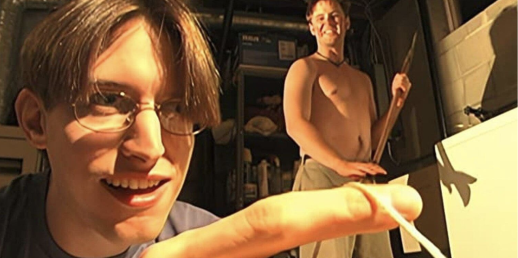 Two men stand in a basement. A man wearing glasses is standing close to the camera; a rubber band is wrapped around his finger. The man behind him is shirtless and holding an axe.