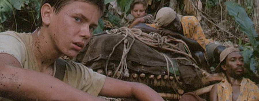 Two children and two men sit in a jungle with a large covered object. The children are wearing beige shirts, and the adults are wearing yellow button downs with blue and white flowers.