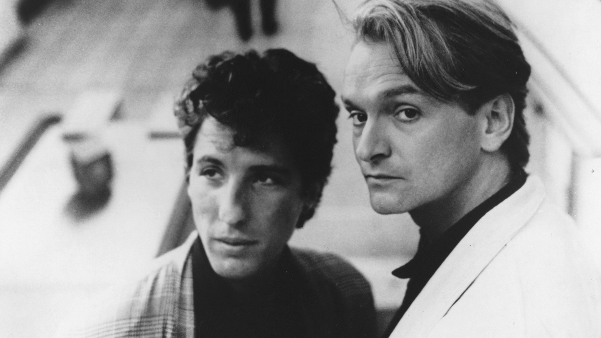 A black-and-white image of Nick Feldman and Jack Hues of the band Wang Chung: two light-skinned young men, one with dark, middle-short curly hair and the other with light, wavy, medium straight hair. Both men are looking off camera in two different directions.