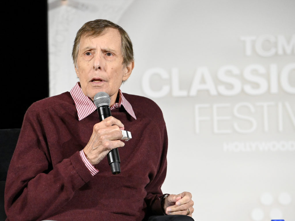 A color image of director William Friedkin speaking into a microphone during an appearance at the TCM Classic Film Festival. He is wearing a maroon sweater over a red-and-white striped shirt and looking toward the audience