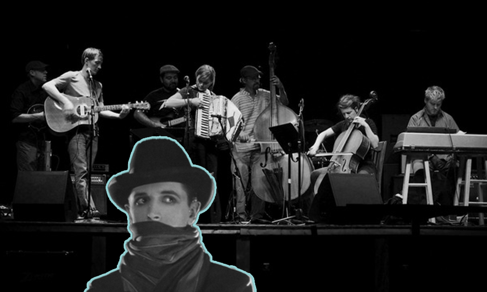 A black-and-white wide-angle shot of seven musicians on stage playing mandolin, guitars acoustic and electric, accordion, upright bass, cello, and keyboard. Superimposed on the image is a man wearing a trilby hat with darting eyes and a scarf concealing the lower half of his face.