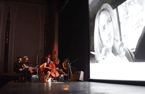 A long shot color photo of the Poor Nobodys as a six-piece dressed in black playing various string instruments in the dark on the left as some members gaze up at the black-and-white film Panacea projected on a large screen on the right, wherein a woman with light hair looks curiously out of the window of a car.