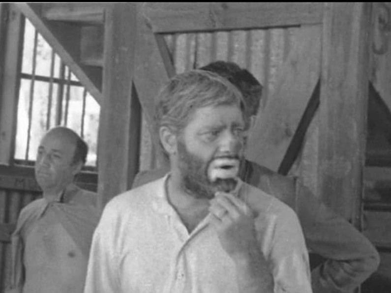 Jerry Lewis wears a white shirt, beard, and clown makeup. He looks to the right, touching his chin thoughtfully. Behind and to the left a bare-chested man stands under a staircase.