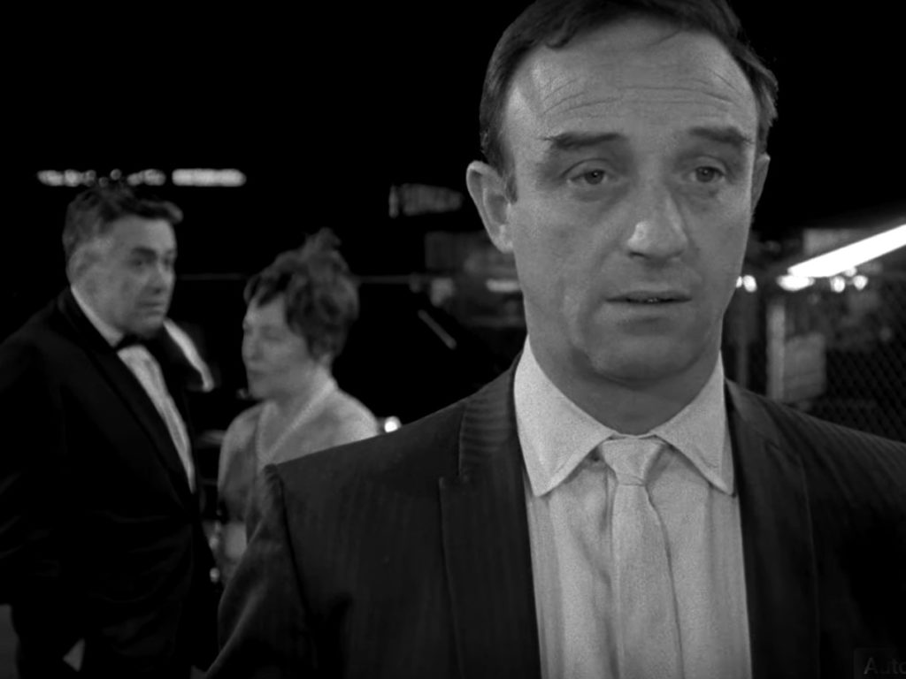 In the foreground, Harry Plotnik wears a suit and tie and a confused expression while two people converse in the background.