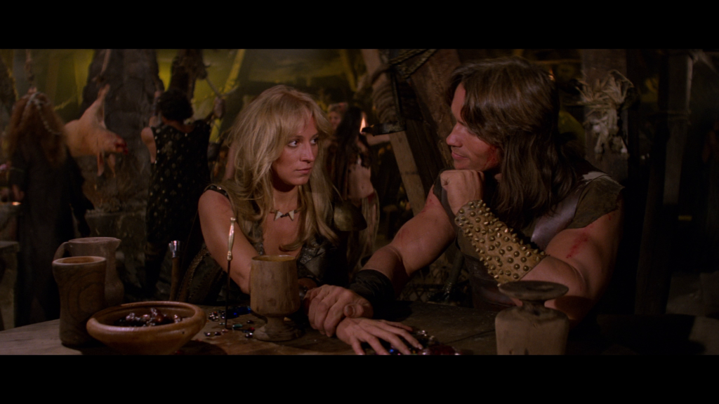 Conan and Valeria are seated together at a tavern. Valeria’s hand is outstretched on the table, with Conan’s hand on her wrist. (The guy has a sensitive side.)