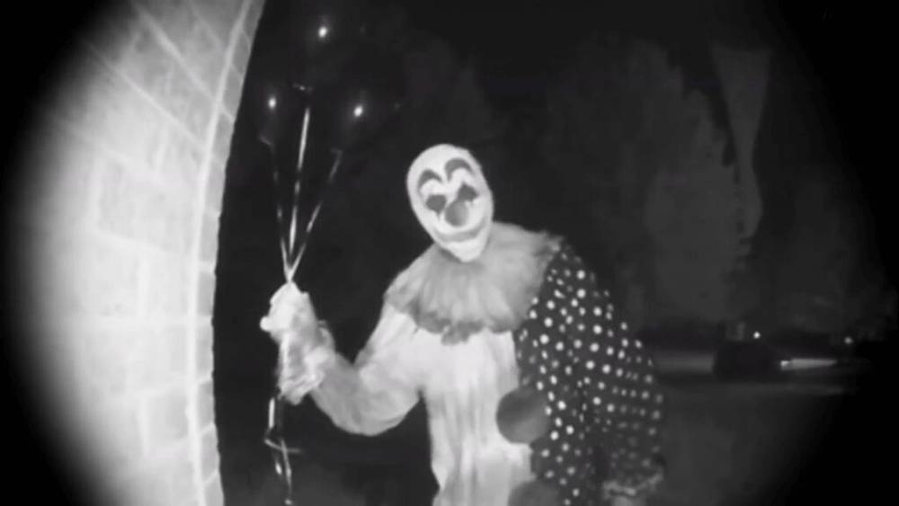 A blurry black and white image by a door camera, showing a person dressed as a scary clown, holding three balloons, standing in someone's doorway, facing the camera.