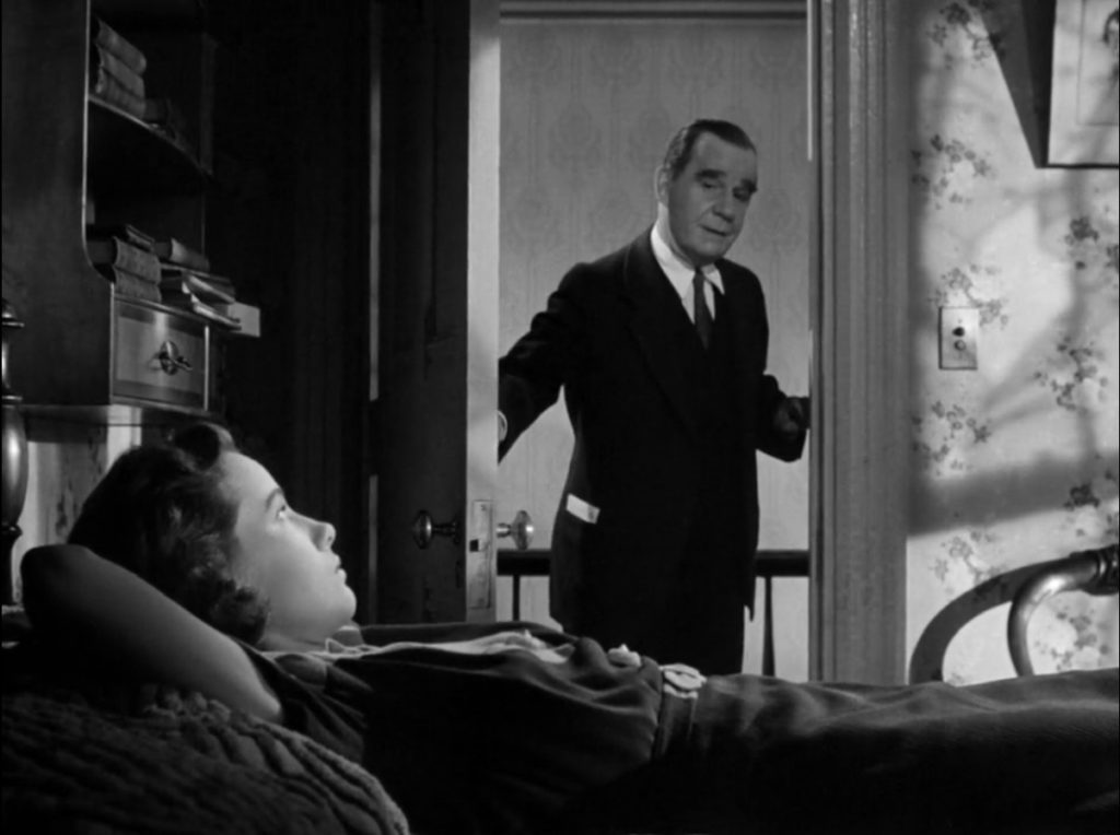 A young woman lays in bed with a bored expression as her father stands in the doorway