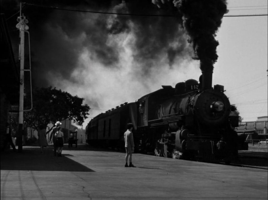 A train puffing black smoke pulls into a station while a child on a platform stands in its shadow