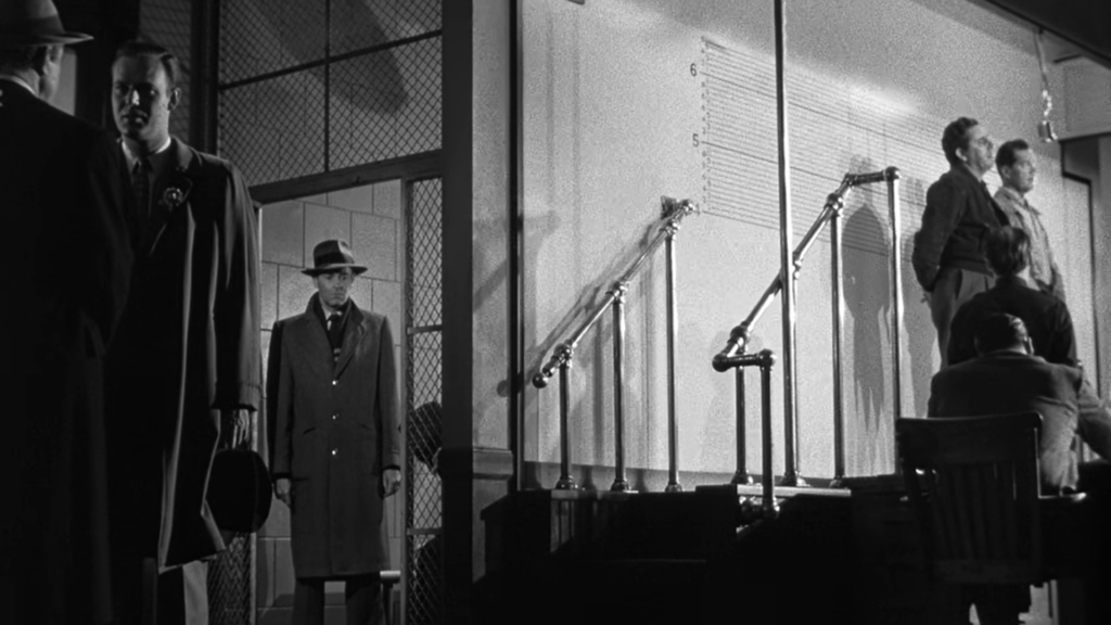 A man in a fedora and a coat walks into a dimly lit room from the back left. There are other men posing in the upper right for a mug shot.