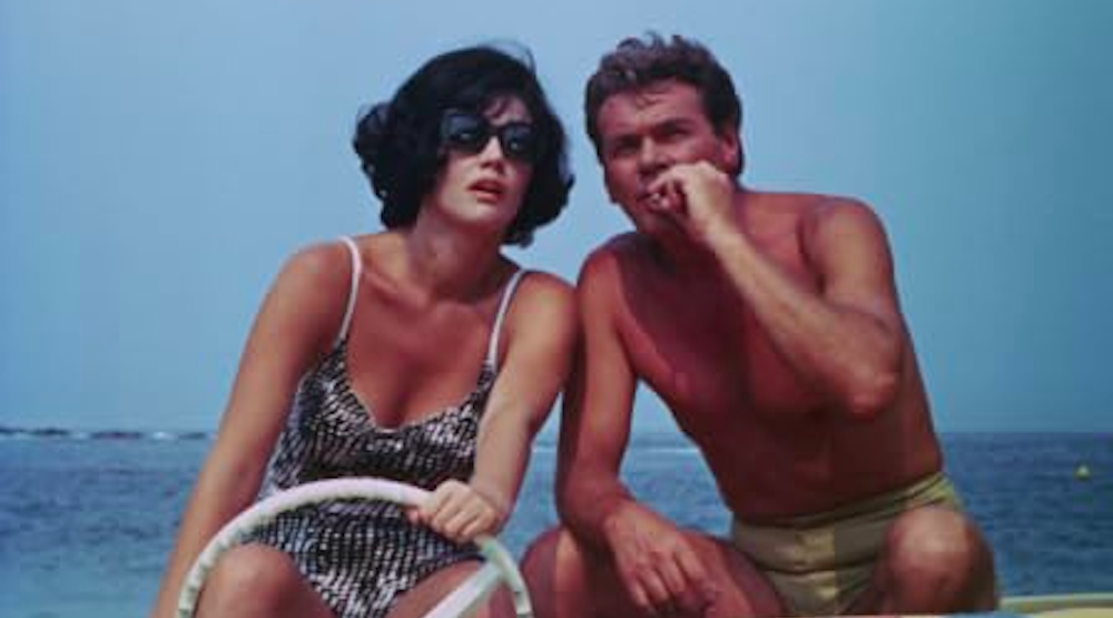 a tan woman with short black hair sitting behind the wheel of a boat next to a tan man with light brown hair smoking a cigarette