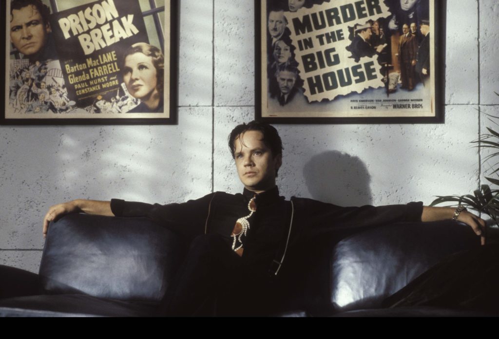 Griffin Mill (Tim Robbins) sitting with his arms spread across a leather couch and posters of the films Murder In the Big House and Prison Break displayed above.