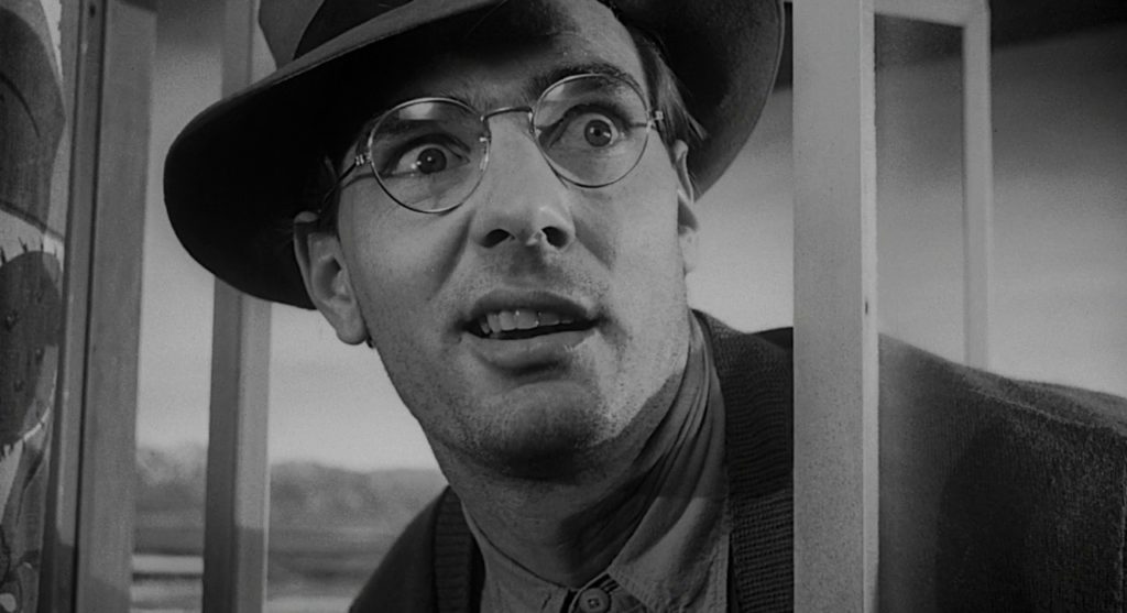 Goofy looking man wearing a fedora hat and wire rimmed glasses pokes his head through a window.