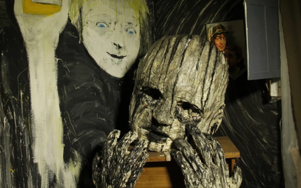 In a black painted room is a large paper mache baby head with black spots for eyes and black paint pouring down its face. Behind it is a blonde woman with blue eyes, who is holding up a bottle of honey just off screen.