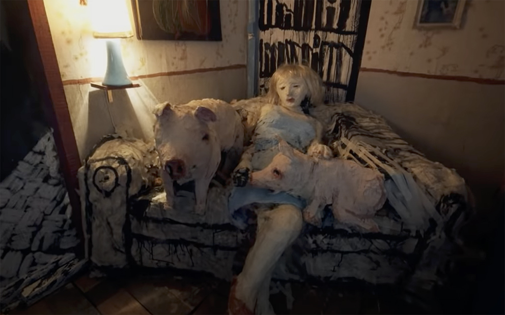 Maria, a paper mache girl with a blonde wig and a blue dress, sits on a white paper mache couch. Two paper mache pigs with haunting black eyes sit on the couch with her.