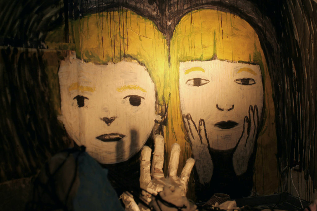 Two blonde children are painted onto a black wall. A boy with short hair is on the left and a girl with long hair is on the right. A paper mache hand is extending from the bottom.