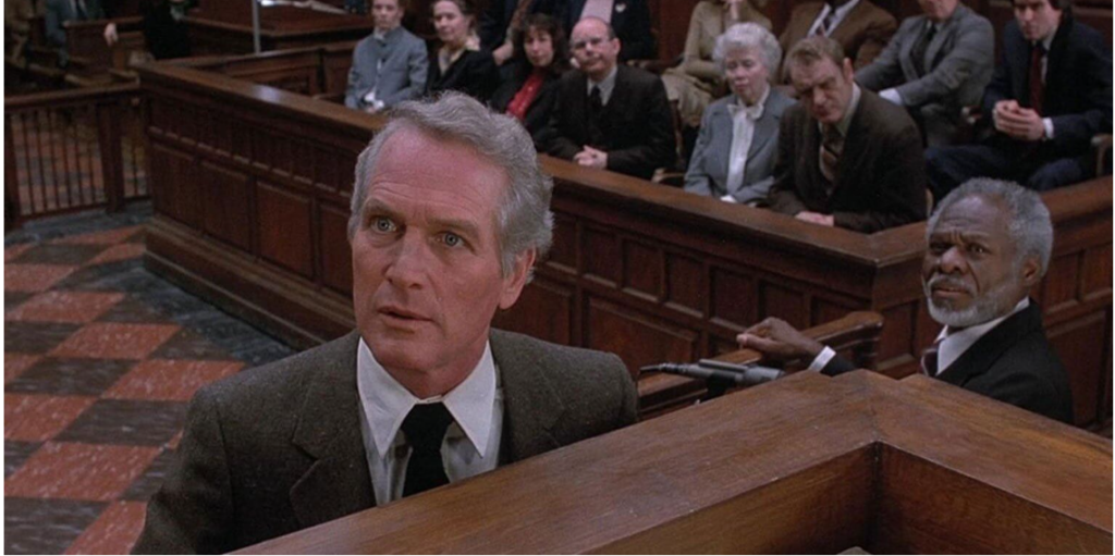 Paul Newman approaches a judge's seat from the left, his right eyebrow cocked in confusion. Directly behind him is the jury; to his right is a witness dressed in a black suit.