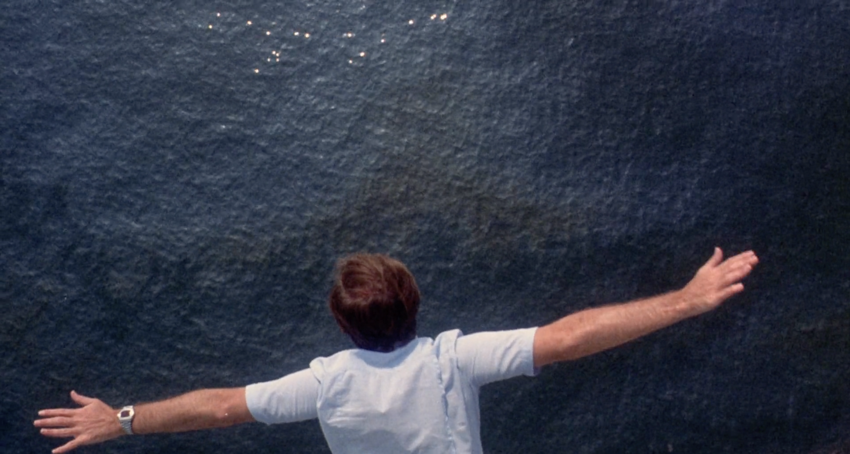 A man in a white t shirt hangs over a calm sea with his arms outstretched. Only his upper body is in the frame. He is facing the water.