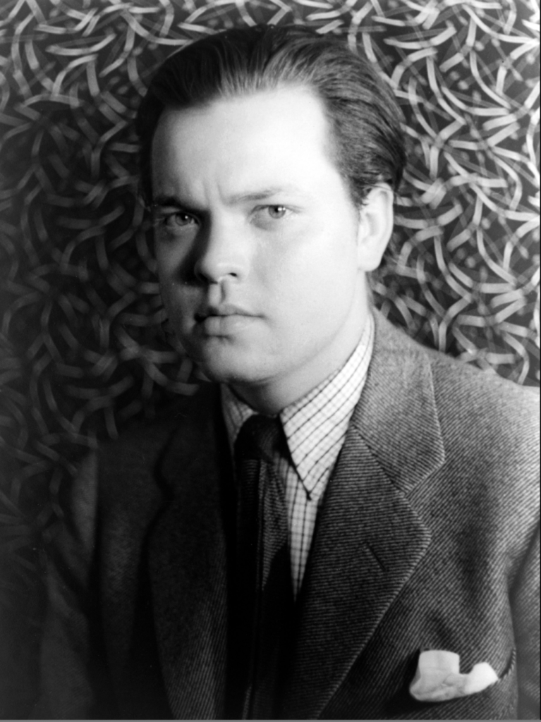 Orson Welles poses in a sharp suit and tie with a plaid button down underneath. The photo is in black and white.