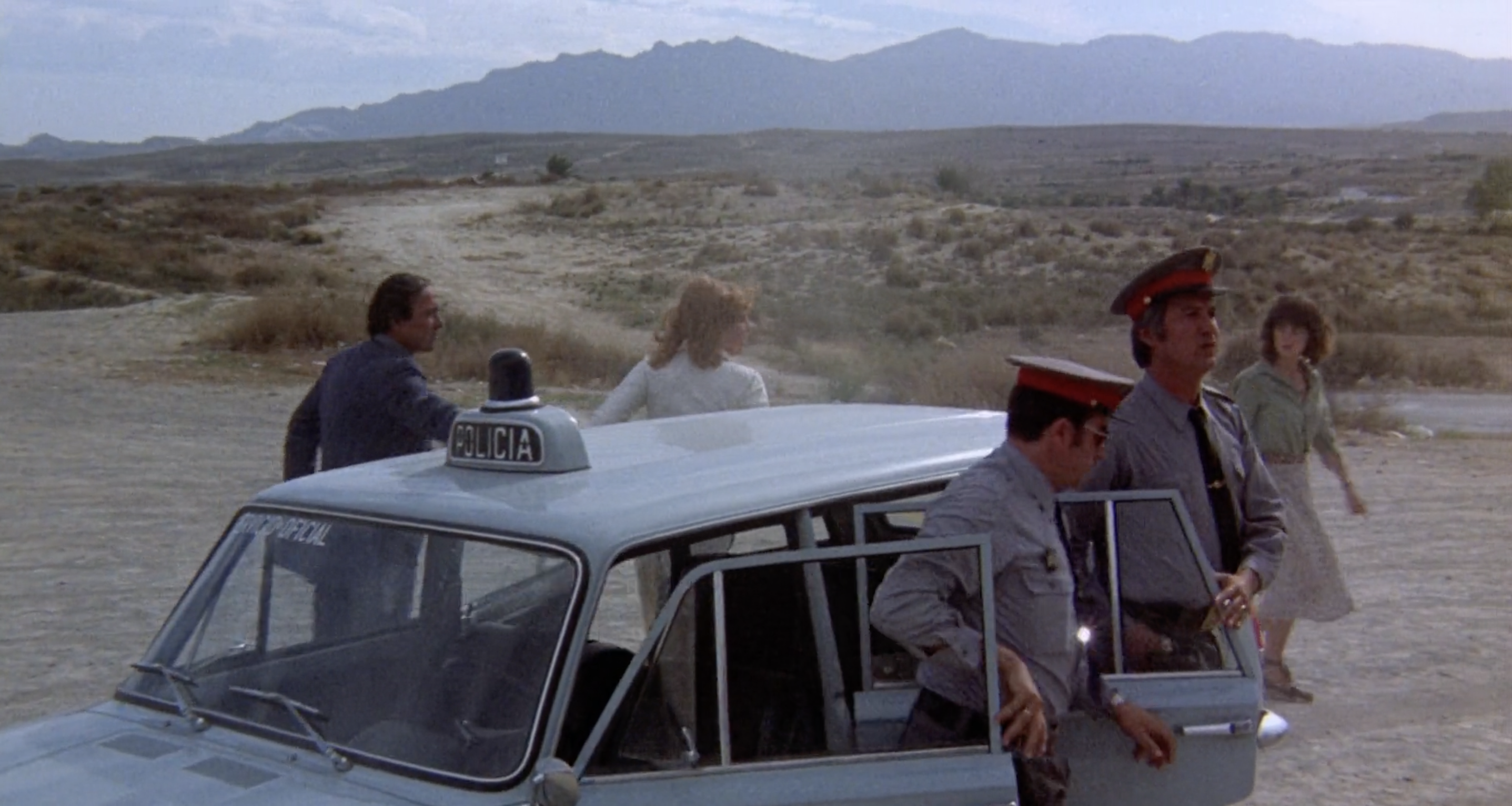 A woman in a green blouse walks across the screen from left to right in the background. In the foreground a woman with a white shirt gets out of a police car along with three policemen.