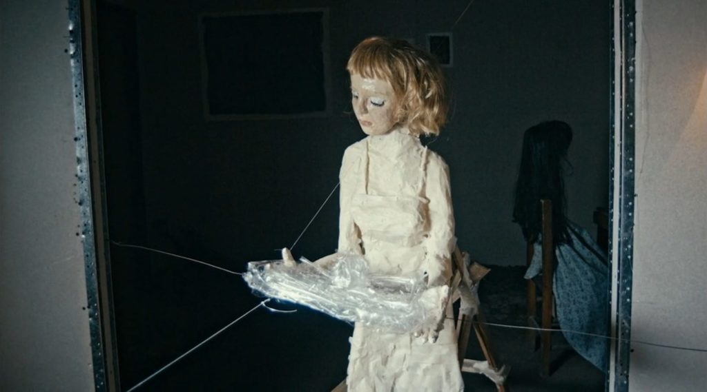 A blonde girl with a beige masking tape dress enters a room carrying a tray made of clear tape.