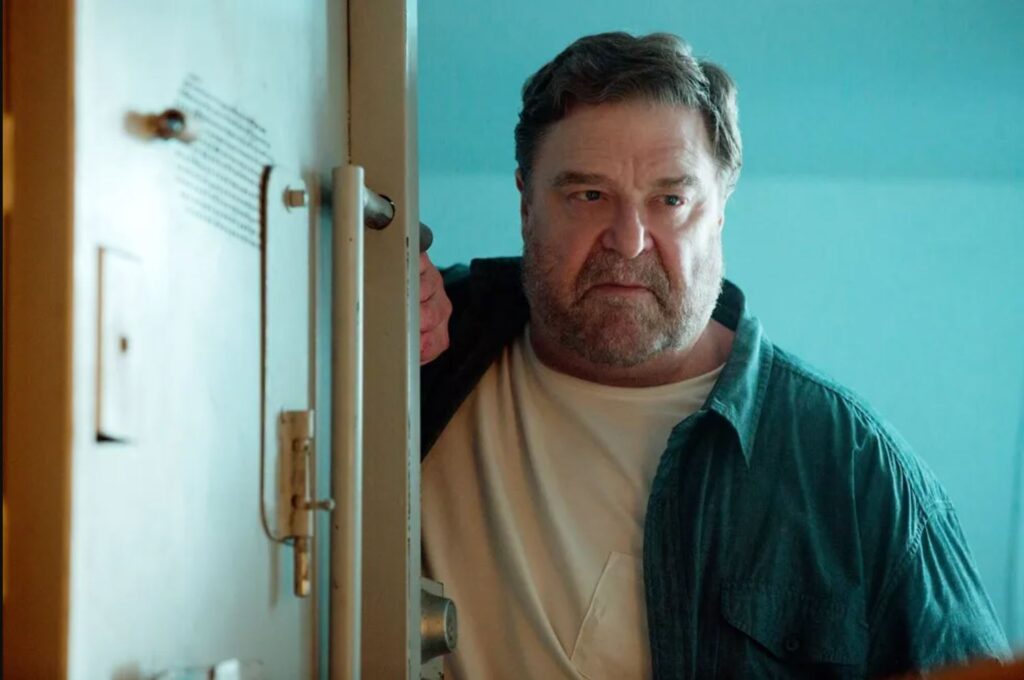 (large man with a grey and brown beard wearing a blue button down and white t-shirt looks menacingly, with a slight frown and squinted eyes, while holding open an industrial door