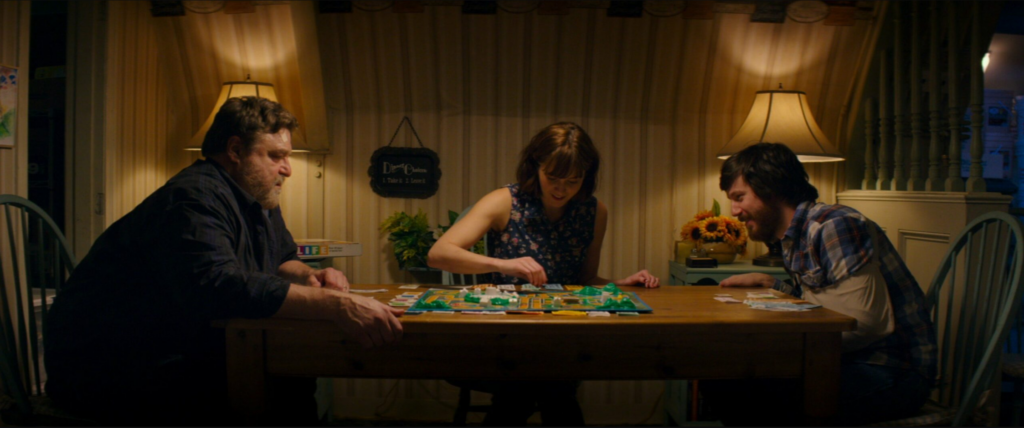 (3 people on 3 different sides of a table playing a board game while laughing and smiling. One is a large man in a blue button-down and jeans at the head of the table, at the other end is a younger man wearing a cast and plaid button-down, and in the middle is a woman wearing a floral print button-down tank top.