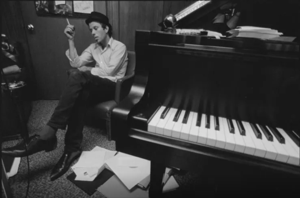 Tom sits cross-legged in a chair, holding a cigarette, piles of music sheets at his feet, a large piano dwarfing him in the foreground, Hollywood, American Zoetrope Studios
