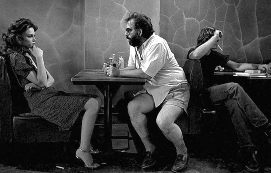 A bearded Coppola in a button-down shirt and shorts, sits in a booth across from actress Diane Lane, giving her direction on the set of “Rumble Fish"