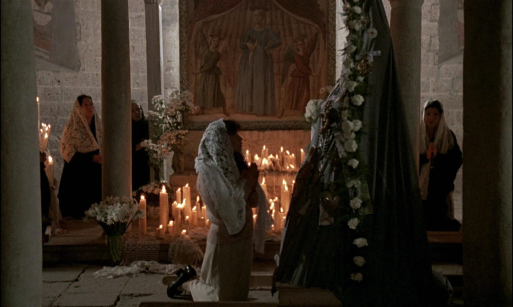 A young woman in a church kneels in front of what seems to be a religious icon—perhaps a representation of Mother Mary. Behind the religious adherent, there’s an altar covered with lit candles, and other women kneeling and observing the ceremony.
