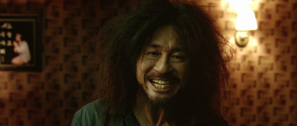 A man with large and unkempt hair (Oh Dae-Su) stares towards the viewer grimacing through a disturbingly large smile.