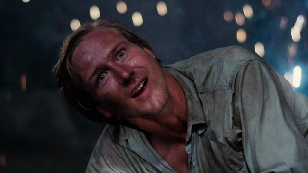 Sweaty man in a dirty shirt stares wide-eyed at the camera as soft lights flicker in the night sky behind him.