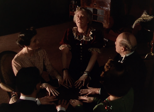 Margarite Rutherford as Madame Arcati leading the group in a seance by having all four dinner guests put their hands in a circle with their fingers touching. The guests are all light skinned, wearing formal dinner party attire- the men in tuxedos, the women in gowns from the scene in Lean’s 1945 film version of Blithe Spirit.