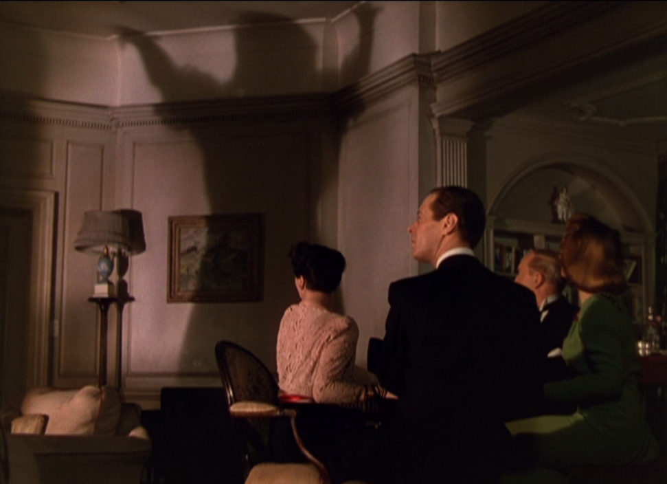 Madame Arcati’s shadow looms on the wall over the the four light-skinned, upper-class dinner guests from the 1945 film version of Blithe Spirit.