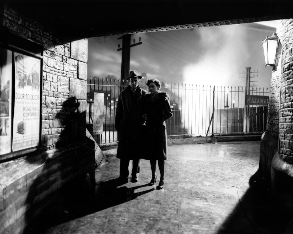 A light-skinned man with a hat and a light-skinned woman, both wearing trench coats, are standing in the archway of an overpass; a metal fence and electric poles linger in the background. The black-and-white image magnifies their shadows on the overpass wall