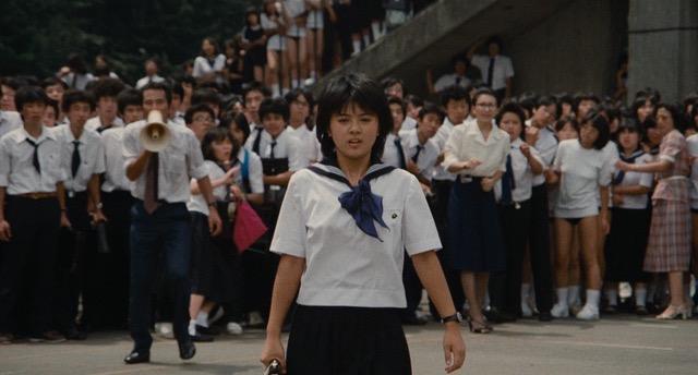 Still from the film, Izumi in the foreground looking straight ahead, with a crowd of classmates in the background. 