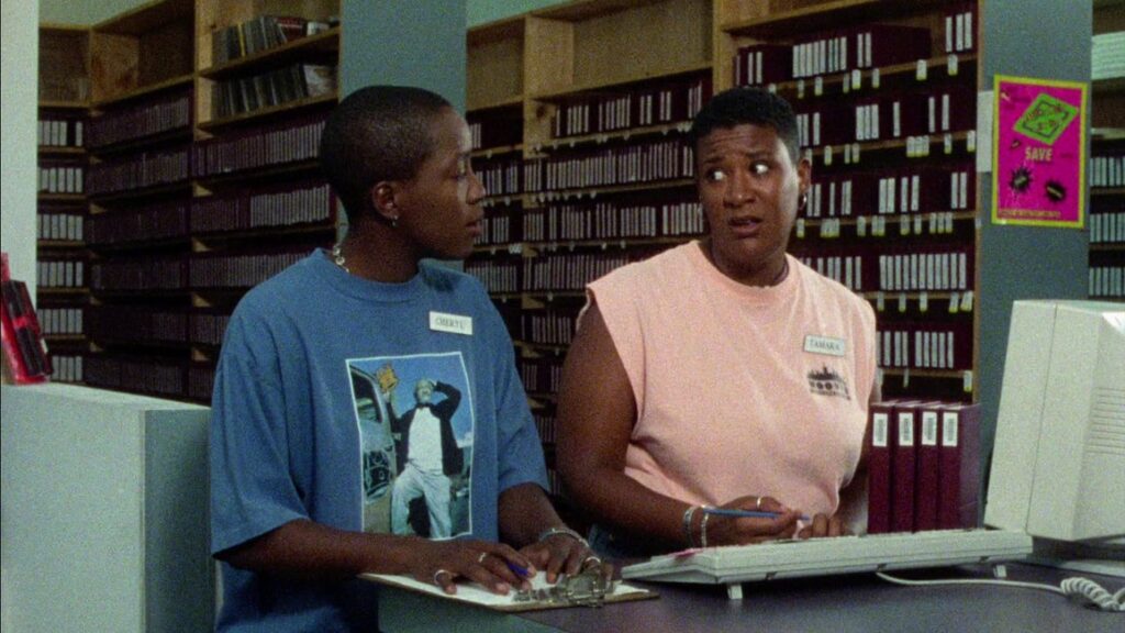 Cheryl, who wears an oversized blue tee shirt and her employee name tag, stands behind the video rental store counter with Tamara, who wears a peach muscle tee and her own name tag. Tamara looks put out by what Cheryl has just told her.