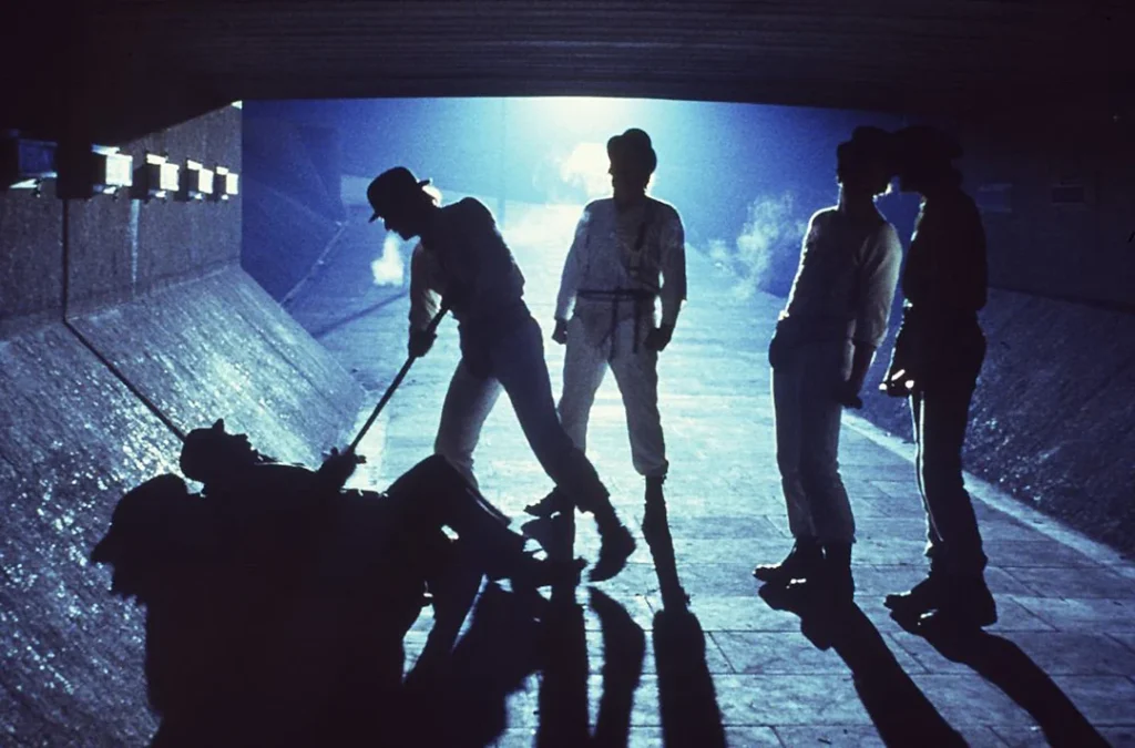 A still from A Clockwork Orange. Alex DeLarge presses a cane into the stomach of a prone man while Alex’s three friends look on.