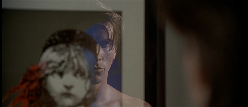 A still from American Psycho. Patrick Bateman looks at his reflection in a framed Les Miserables poster.