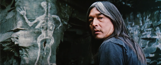 Medium shot of Graham Greene as Arthur right of center in frame with cliffs featuring Indigenous paintings, including one prominent semi-amorphous figure. Arthur is looking over his left shoulder at the camera wearing a black t-shirt and sporting a long mane of dark silver-tinged hair. The First Nations character has an expression of resignation and inscrutability on his face.