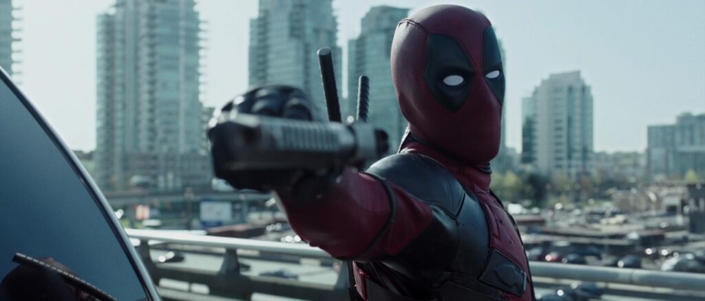 A still from Deadpool. Deadpool wears a red leather suit that covers his face and body and points a handgun at the camera.