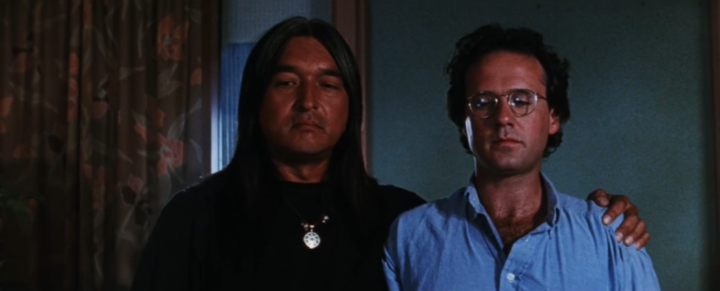 Two men stand together in a hotel room with teal walls and a brown curtain with red flowers hanging on the left side of the room. One man has long black hair and is wearing a black shirt, the other has a blue shirt and short brown hair. He is wearing glasses.