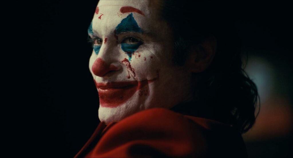 A still from Joker. Arthur Fleck wears clown makeup on his face with a splatter of blood and smiles menacingly.