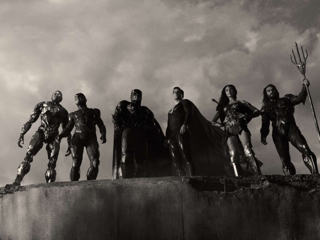 A still from Zack Snyder’s Justice League. The Justice League heroes stand against a cloudy sky in black in white.