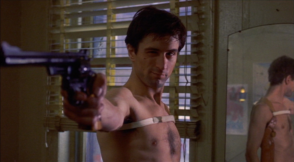 A still from Taxi Driver. A shirtless Travis Bickle points a large handgun toward the camera