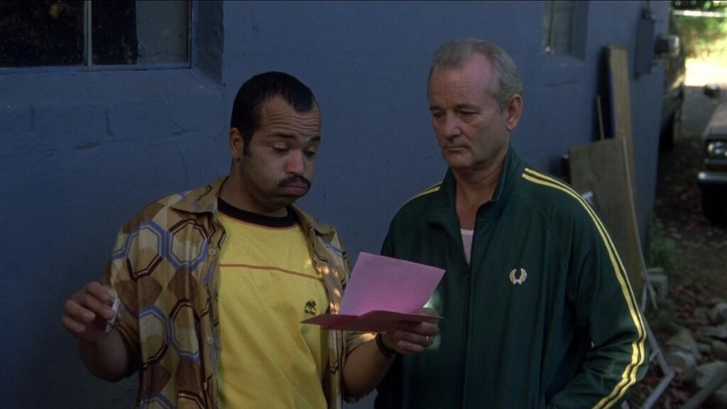 Winston holds the letter and a joint. He wears a retro collared shirt over a yellow tee. He puffs out his cheeks and raises his eyebrows, looking surprised and baffled. He stands against a blue wall next to Don, who wears a green tracksuit and looks at the letter with a slightly mournful expression.