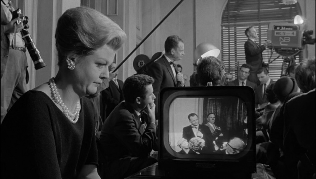 A woman, Eleanor Iselin, watches her senator husband, John Iselin, on a television screen as he creates chaos in a press conference