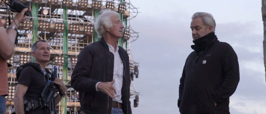  Roger Deakins stands to the left of Sam Mendes. Deakins is a light-skinned man in his seventies and has white hair, a black jacket and a white shirt. Sam Mendes is in his fifties; he has a medium-light skin tone and gray hair and has his chin tucked into the collar of his black jacket. Nearby stand two members of the crew. Behind them is a tall structure made of scaffolding with dozens of groups of large round lights mounted on it.