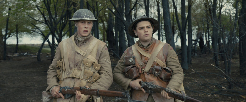 chofield and Blake are young men with light skin tones. They wear period-appropriate khakhi-colored uniforms and metal helmets and both carry rifles. Blake is saying something to Schofield, who looks dubious. Behind them is a small copse of trees.