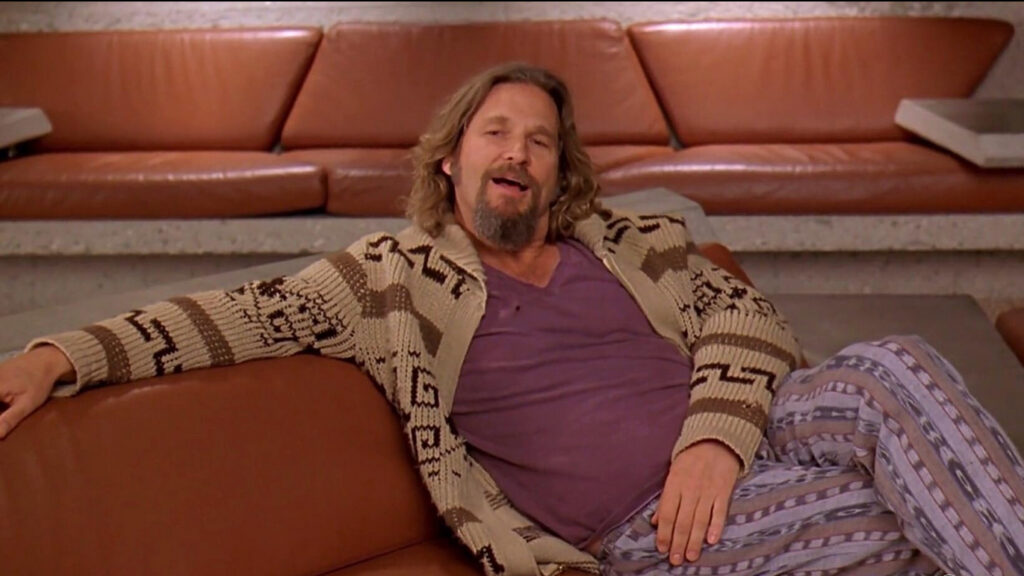 The Dude sprawls on a leather couch wearing his signature Guatemalan print pants and cardigan over a stained tee shirt. His relaxed pose and expression suggest he is overconfident.